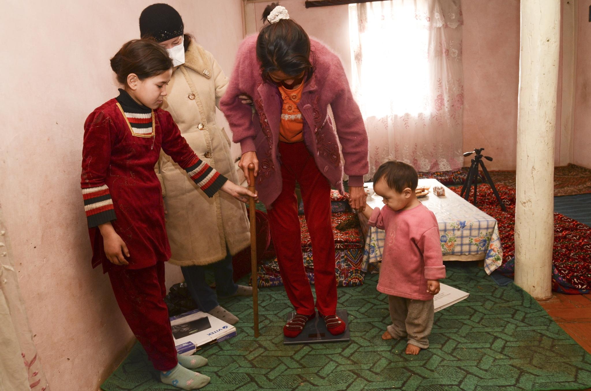 A filha Nadira e o neto Atabek ajudam Rohatay a subir na balança. A enfermeira da comunidade vai todos os dias até a casa da paciente para levar a dose diária de medicamentos e os comprimidos contra os efeitos colaterais e também para garantir que a paciente está fazendo o tratamento. Sayyora controla o peso da paciente e os novos sintomas de efeitos colaterais causados pelos medicamentos contra a tuberculose. © Andriy Slizkiy