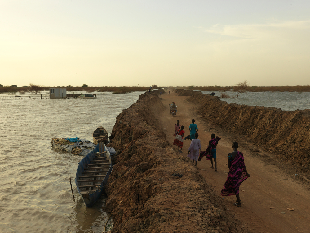 Pessoas caminham ao longo da estrada entre diques inundados ao redor do acampamento de deslocados internos de Bentiu. Foto: Peter Caton, Sudão do Sul, abril de 2022.