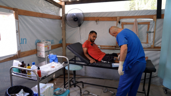 Profissional de MSF examina o ferimento de Saad Hussein, que  vive com um fixador externo após ter sido ferido em 2025 durante uma distribuição de alimentos realizada pela Fundação Humanitária de Gaza. Centro de Atenção Primária à Saúde Al Mawasi, Khan Younis, Gaza. ©Nour Alsaqqa/MSF