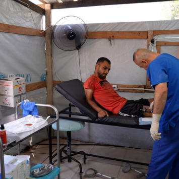 Profissional de MSF examina o ferimento de Saad Hussein, que  vive com um fixador externo após ter sido ferido em 2025 durante uma distribuição de alimentos realizada pela Fundação Humanitária de Gaza. Centro de Atenção Primária à Saúde Al Mawasi, Khan Younis, Gaza. ©Nour Alsaqqa/MSF