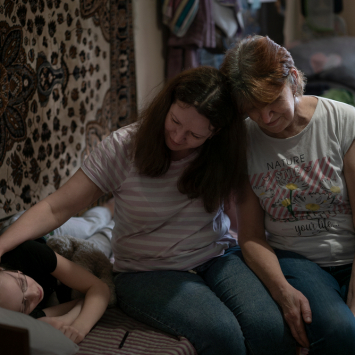 Polina (à esquerda), ao lado de sua mãe, Hanna Bila (centro), e sua avó, Tetiana Yakishina (à direita), no quarto que compartilham no centro de pessoas deslocadas internamente em Vinnytsia, na Ucrânia. © Caroline Thirion/MSF