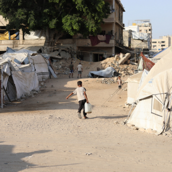 Menino palestino carrega um galão d'água até a tenda onde mora sua família, deslocada pela guerra. Gaza, agosto de 2025. ©Nour Alsaqqa/MSF